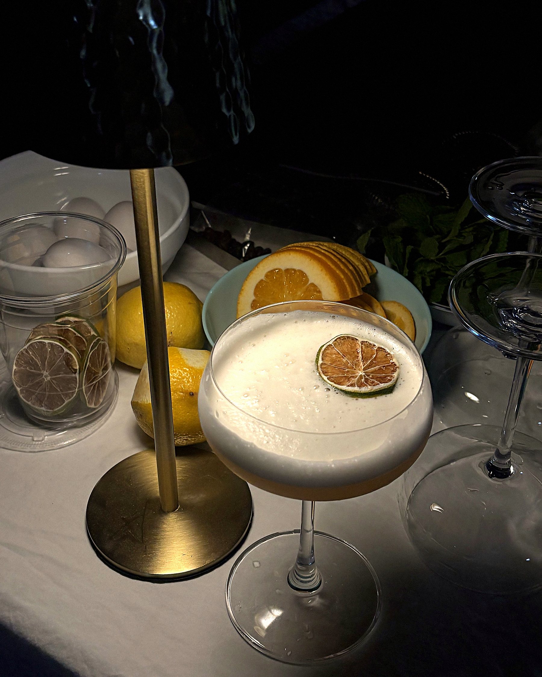 An amaretto sour in a coupe, topped with foam and a dehydrated lime wheel, beside a brass bar lamp